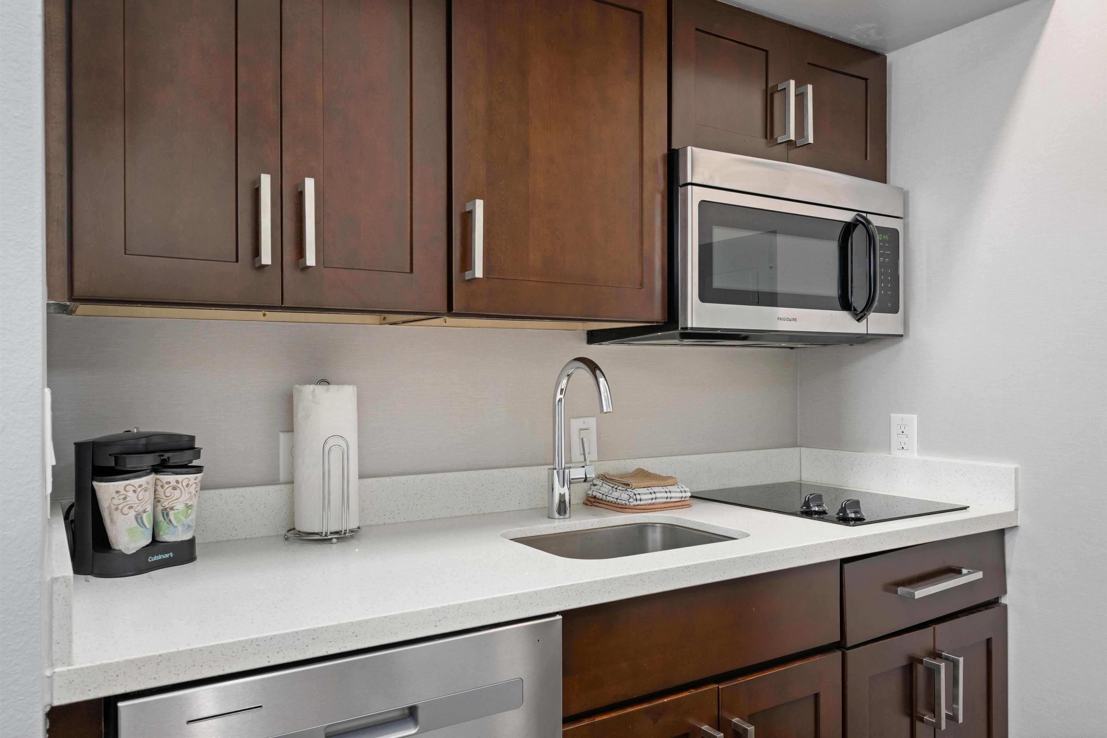 A kitchen with dark wooden cabinets, a stainless steel microwave, a sink, and a dish drying rack.