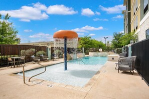 A swimming pool with a water fountain, lounge chairs, and a building in the background. - Homewood Suites By Hilton New Braunfels (New Braunfels)