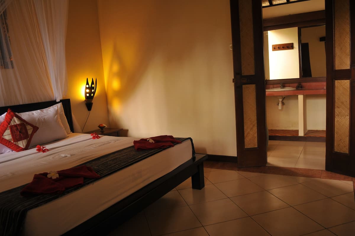 Un lit bien fait, doté d’un baldaquin, une table de chevet avec une lampe, et une vue sur une salle de bain avec un lavabo et un miroir.