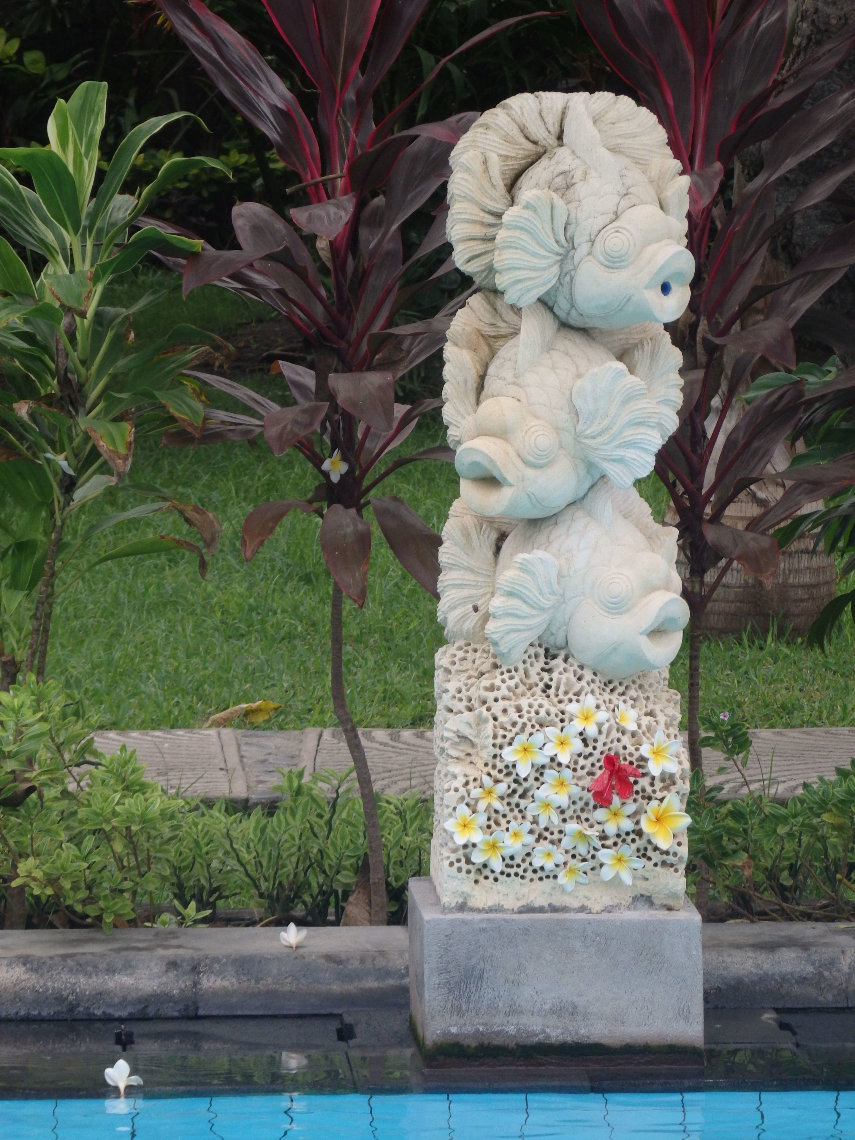 Escultura em pedra de criatura mítica, com base adornada por flores, situada em um jardim com piscina ao primeiro plano.