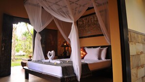 A bed with a canopy, a nightstand with a lamp, and a view of a garden. - Tirta Sari Bungalows & Spa (Pemuteran)