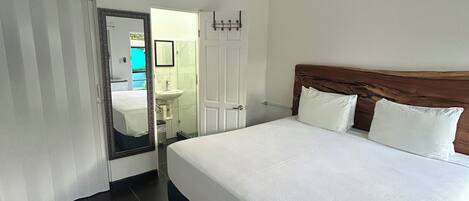 Ein Hotelzimmer mit einem Bett, einem Deckenventilator, einem Badezimmer mit Waschbecken und Spiegel sowie einer Tür zu einem weiteren Raum.