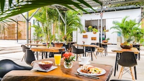 Un café al aire libre con mesas y sillas de madera, rodeado de exuberante follaje y plantas en macetas.