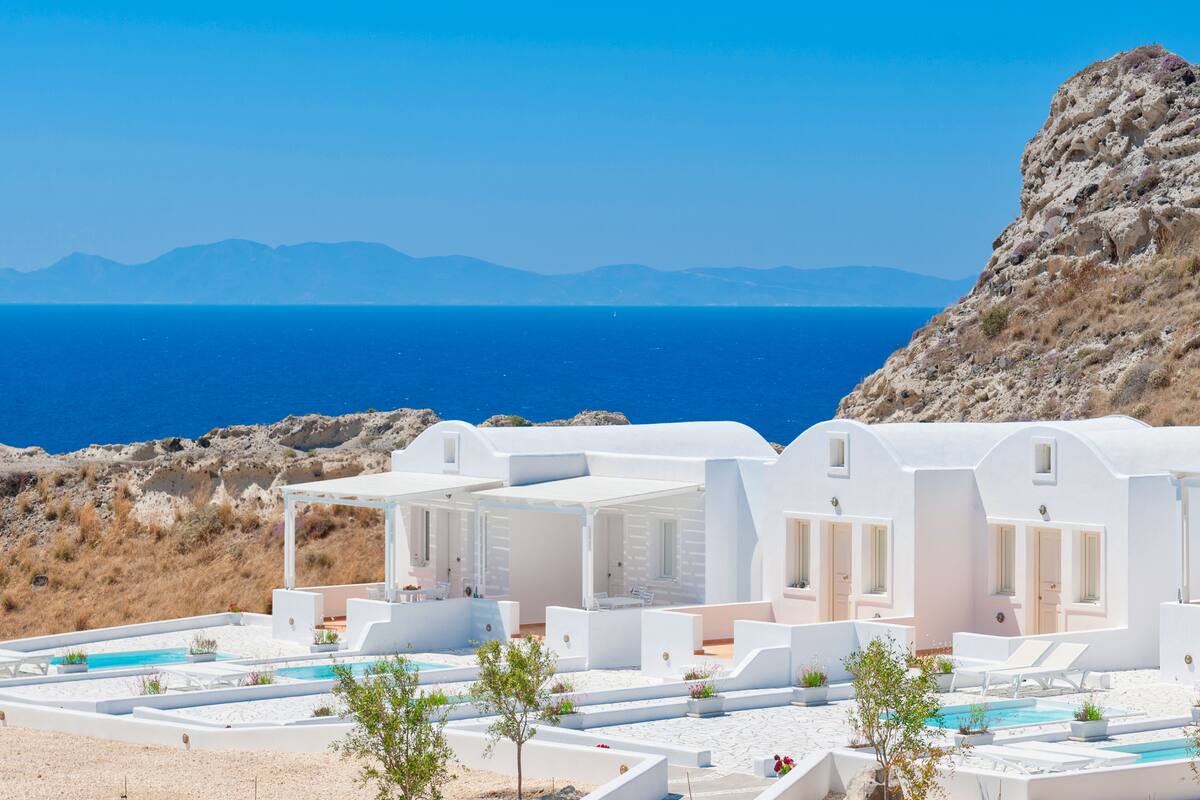 A white building with multiple rooms and a pool area, overlooking a body of water and mountains.