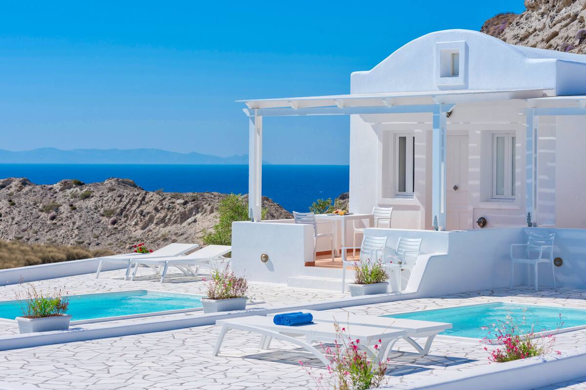 A white villa with a pool and outdoor seating, overlooking a mountainous coastline.
