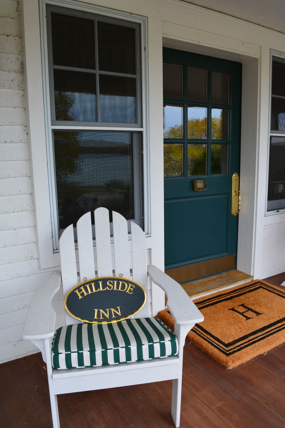 A white Adirondack chair with a green and white striped cushion in front of a teal door with a brass handle.