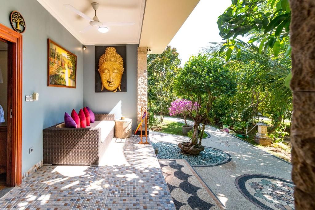 A room with a large Buddha statue, a wicker sofa with cushions, a tiled floor, and a garden with a tree and pebbles.