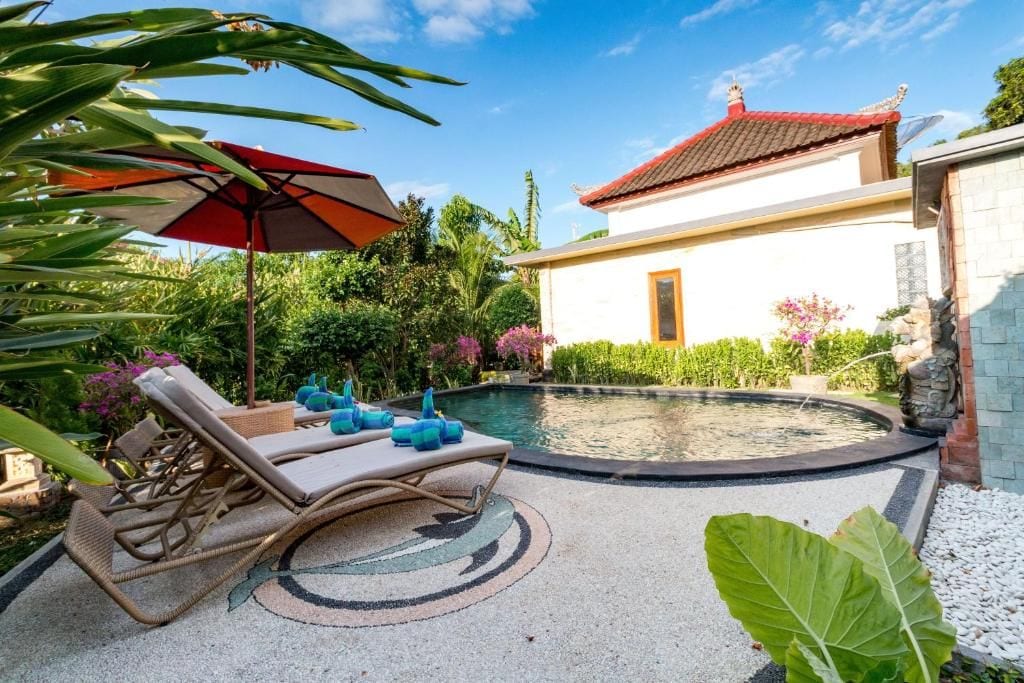 A pool area with lounge chairs and an umbrella, surrounded by greenery and a building with a red roof.