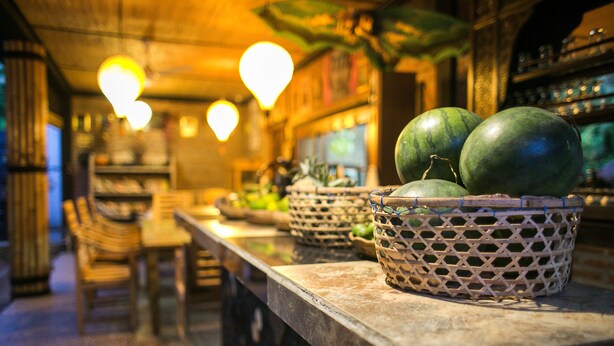 Una zona de bar con cestas de mimbre llenas de frutas, sillas de madera y lámparas colgantes.