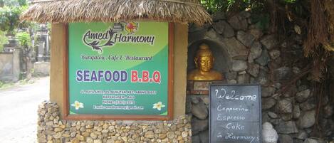 A signboard for Amed Harmony Bungalow, Cafe & Restaurant, featuring a seafood BBQ menu, contact details, and a welcome message for cappuccino, espresso, and cake.