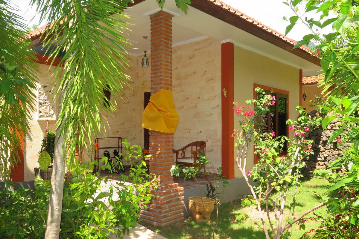 Una casa amb un columnat de maó, un porxo amb cadires i un jardí amb diverses plantes i flors.