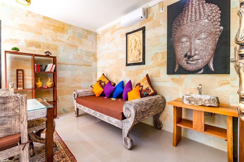 Una habitación con pared de piedra, un sofá con cojines de colores, una mesita de madera, una estantería y una estatua de Buda.