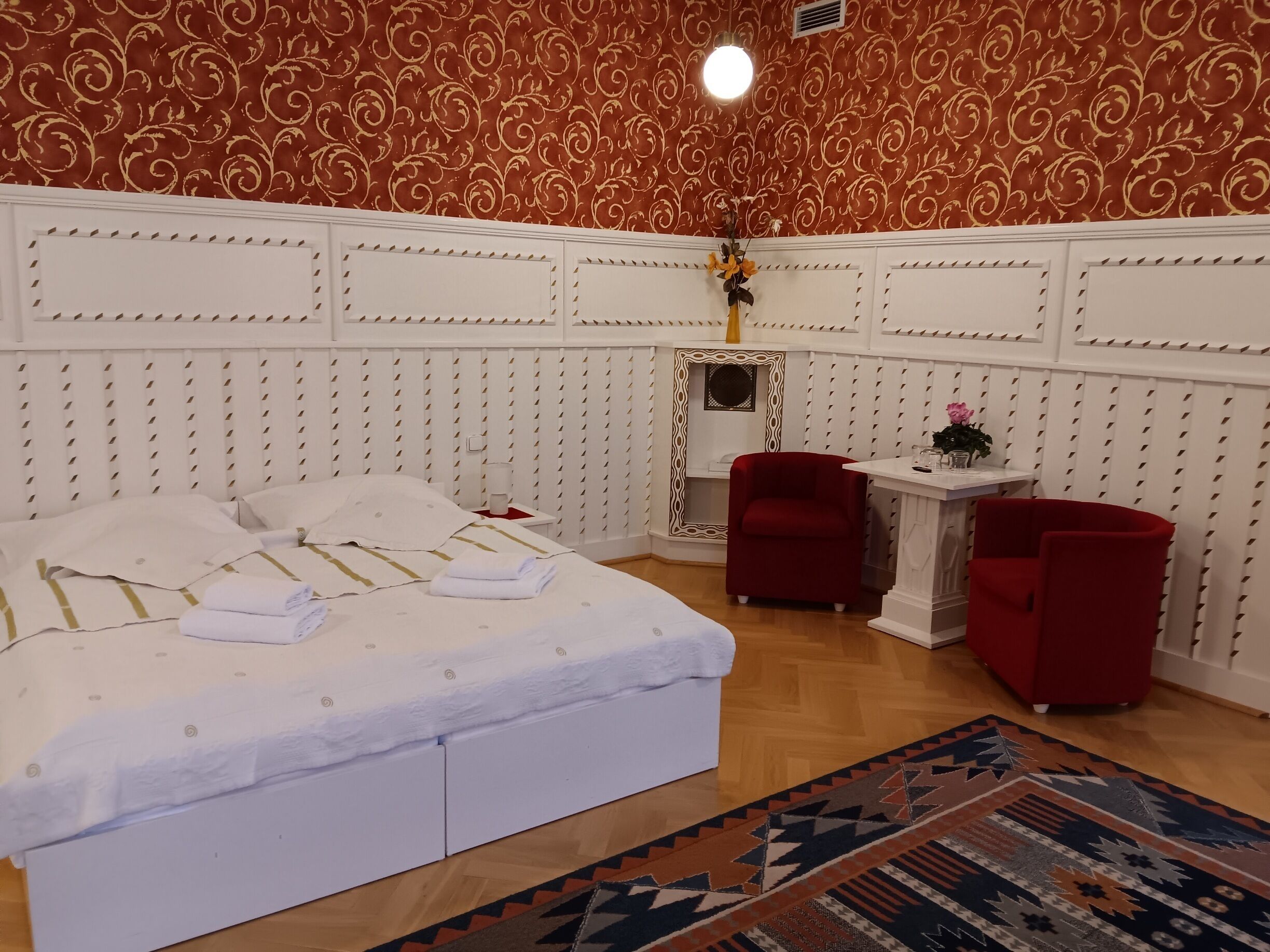 Une chambre d’hôtel avec un grand lit, deux fauteuils rouges, une petite table avec un vase et un papier peint à motifs.