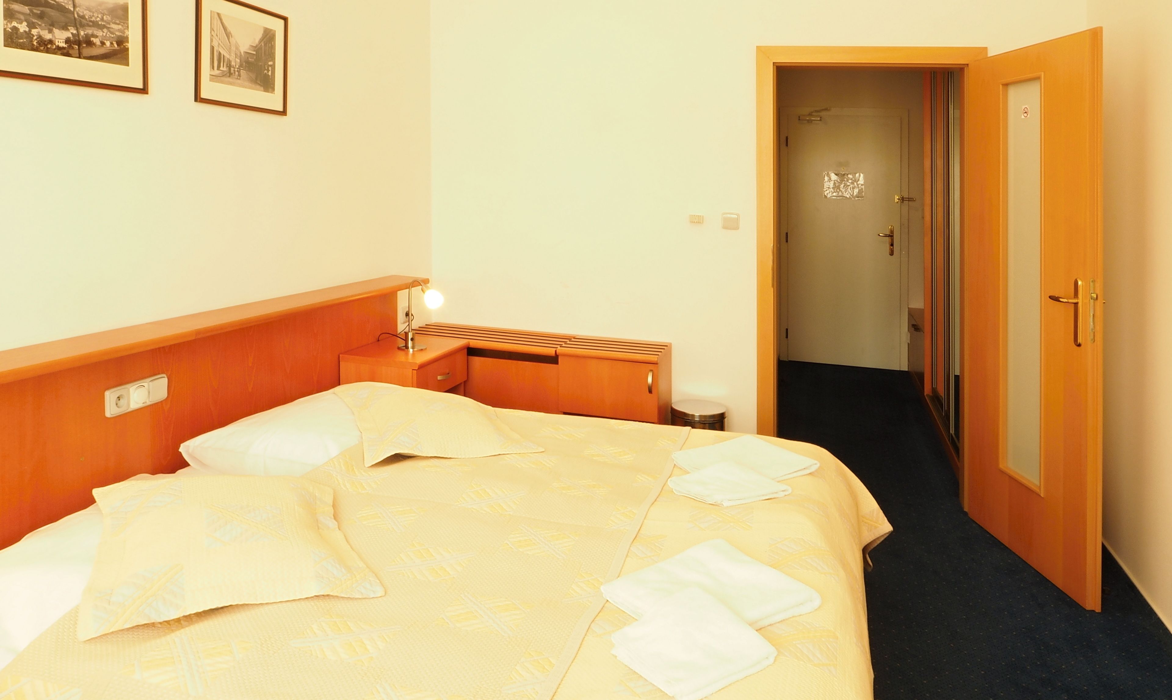 Habitación de hotel con cama, mesita de noche, lámpara y una puerta que da a un pasillo.