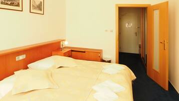 Habitación de hotel con cama, mesita de noche, lámpara y una puerta que da a un pasillo.
