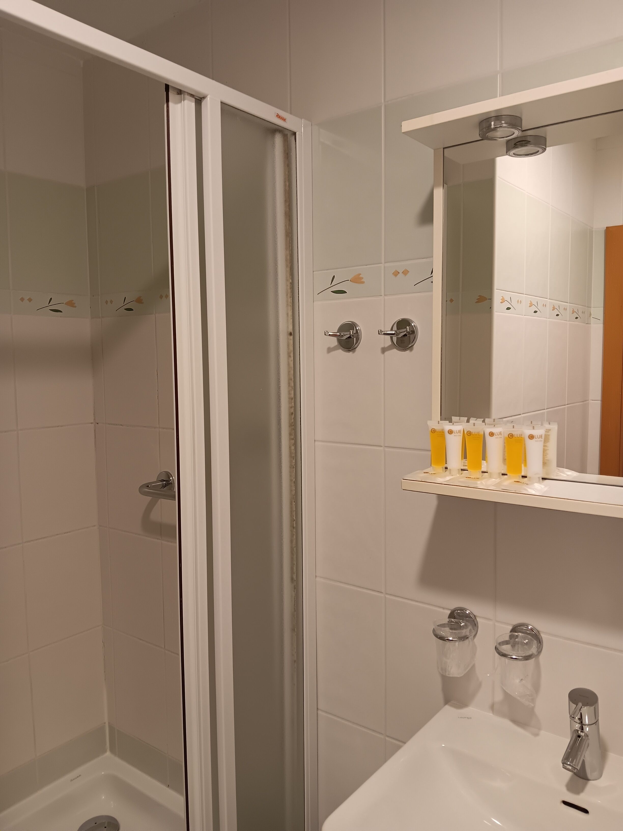 A bathroom with a shower, a sink, a mirror, and toiletries on a shelf.