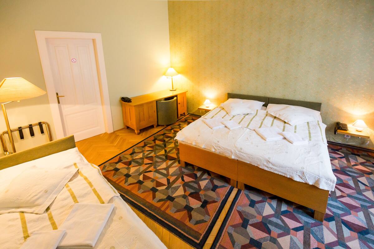 A hotel room with a bed, bedside lamps, a wooden nightstand, a desk, and a patterned rug.