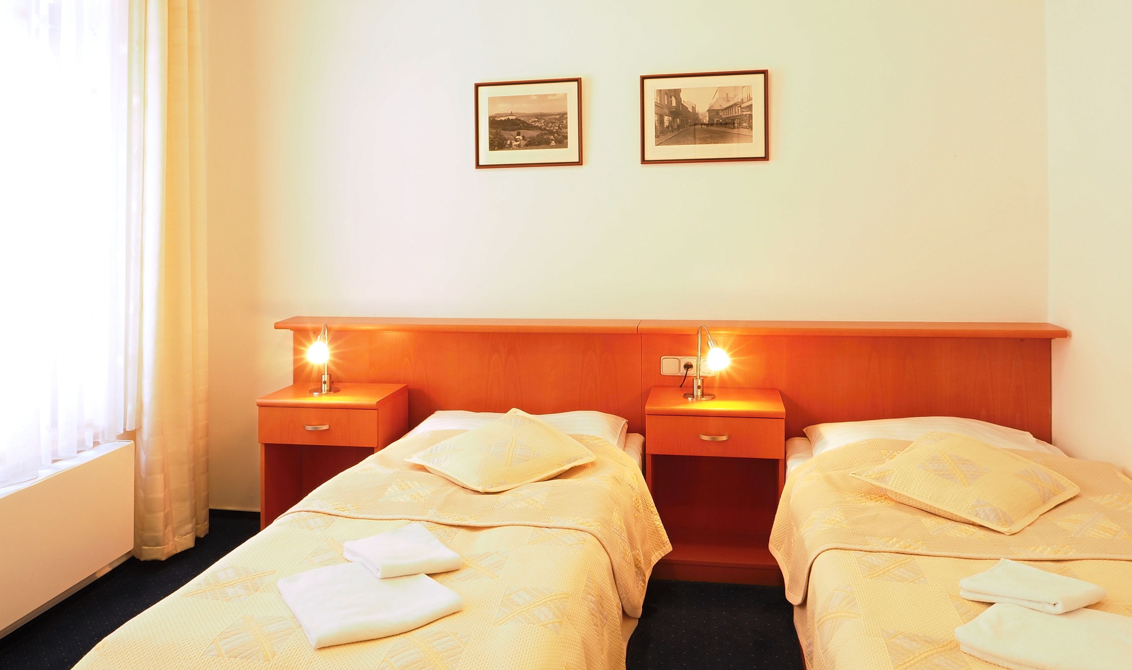 A hotel room with two beds, each with a bedside table and lamp. There are two framed pictures on the wall above the beds.