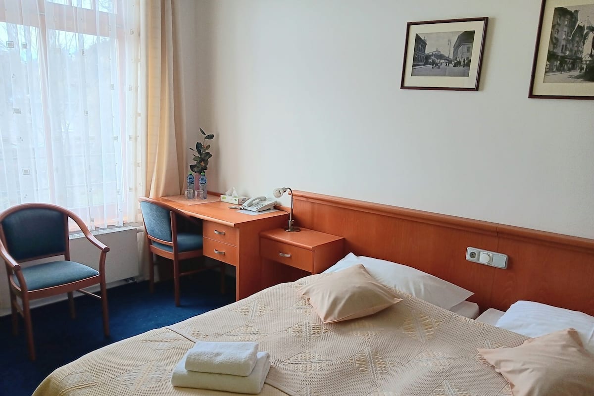 A hotel room with a bed, a desk, a chair, a bedside table, and a window with curtains.