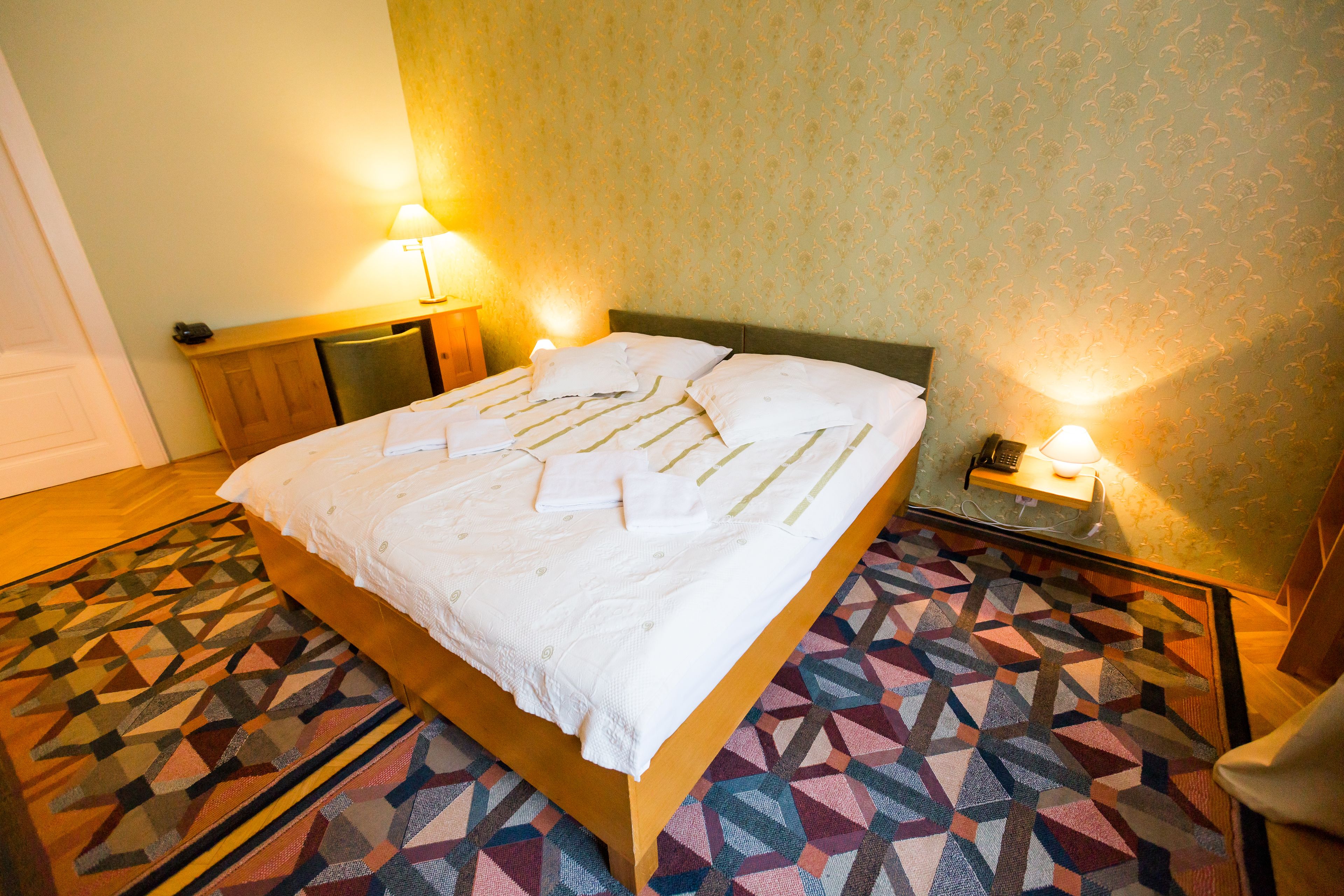 A neatly made bed with a geometric patterned rug, a wooden headboard, and two bedside lamps.