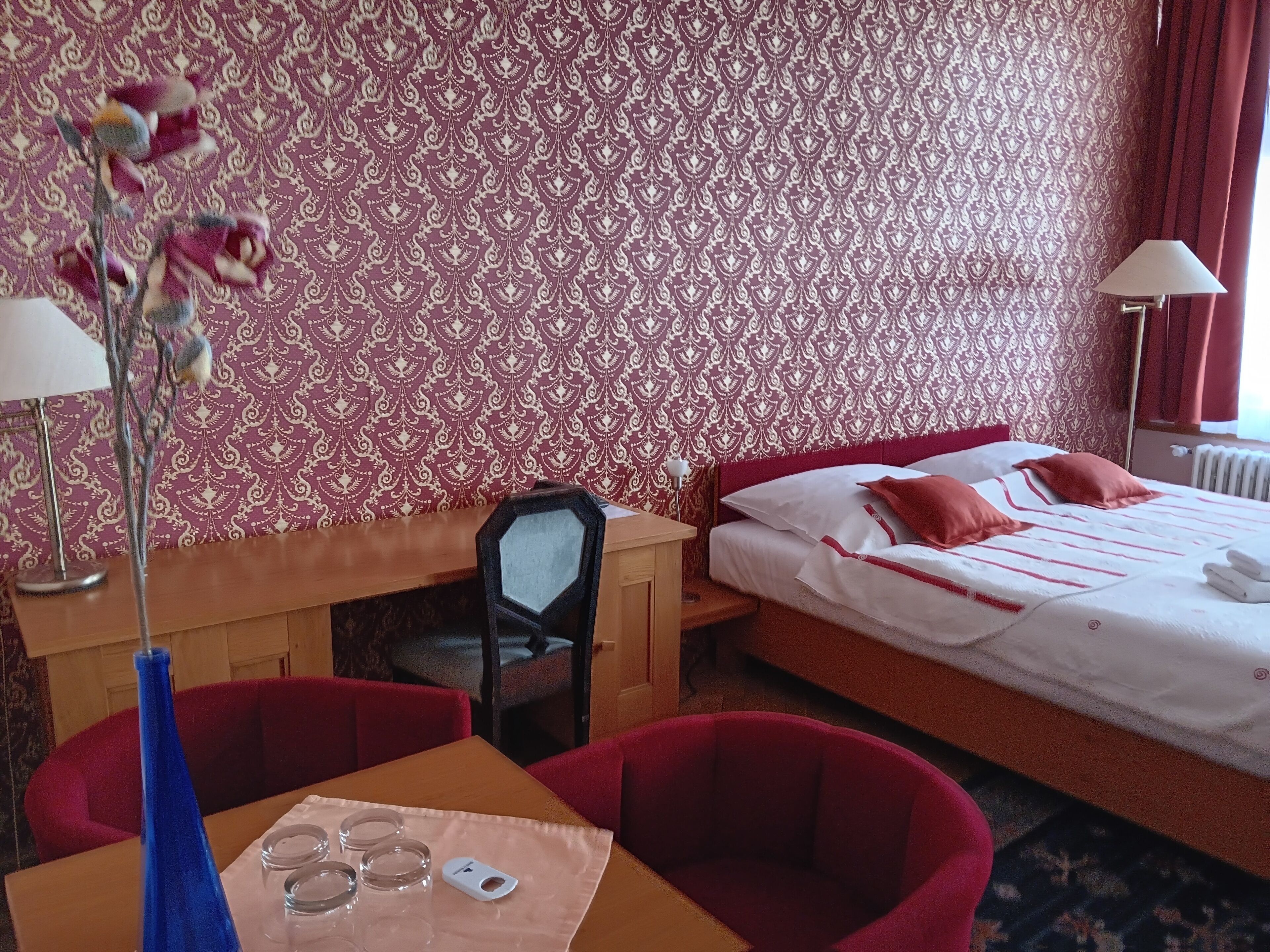 Ett hotellrum med tapet med ett mönster, en säng med röda kuddar, ett skrivbord med en lampa och en stol.