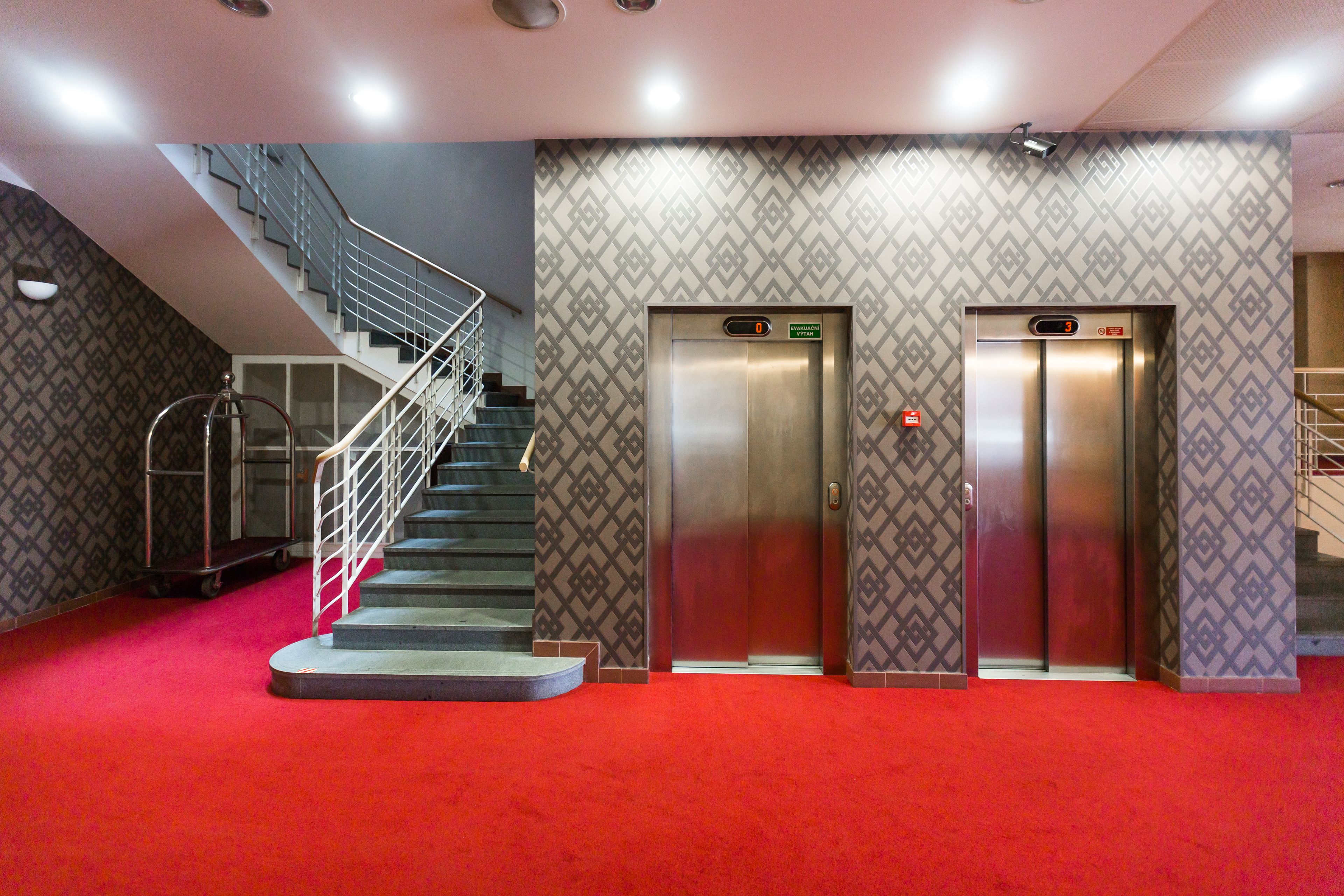 Um saguão de hotel com tapete vermelho, dois elevadores, uma escada e paredes com padrão.