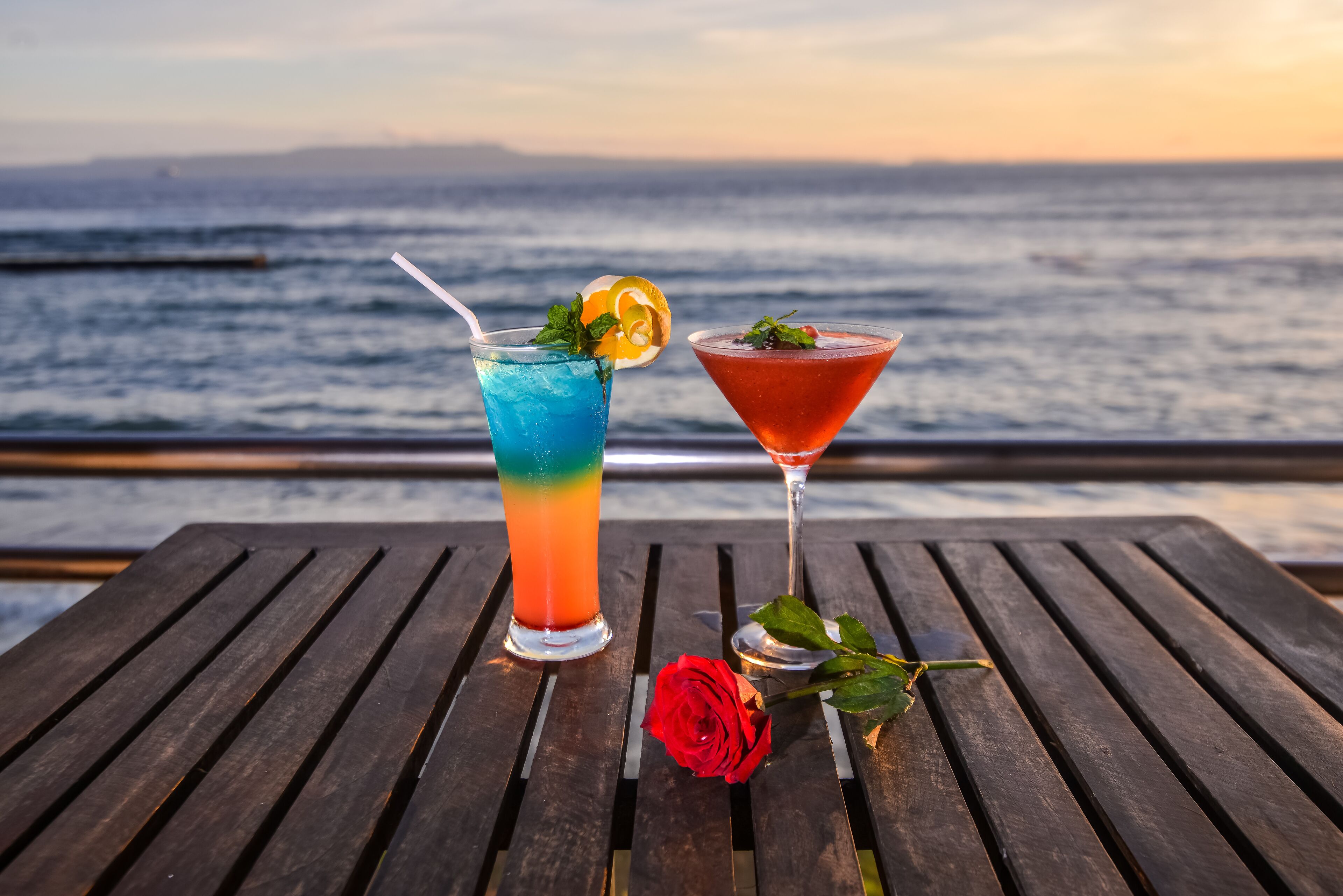Zwei Cocktails auf einem Holztisch, neben ihnen eine Rose, mit Blick auf ein Gewässer.