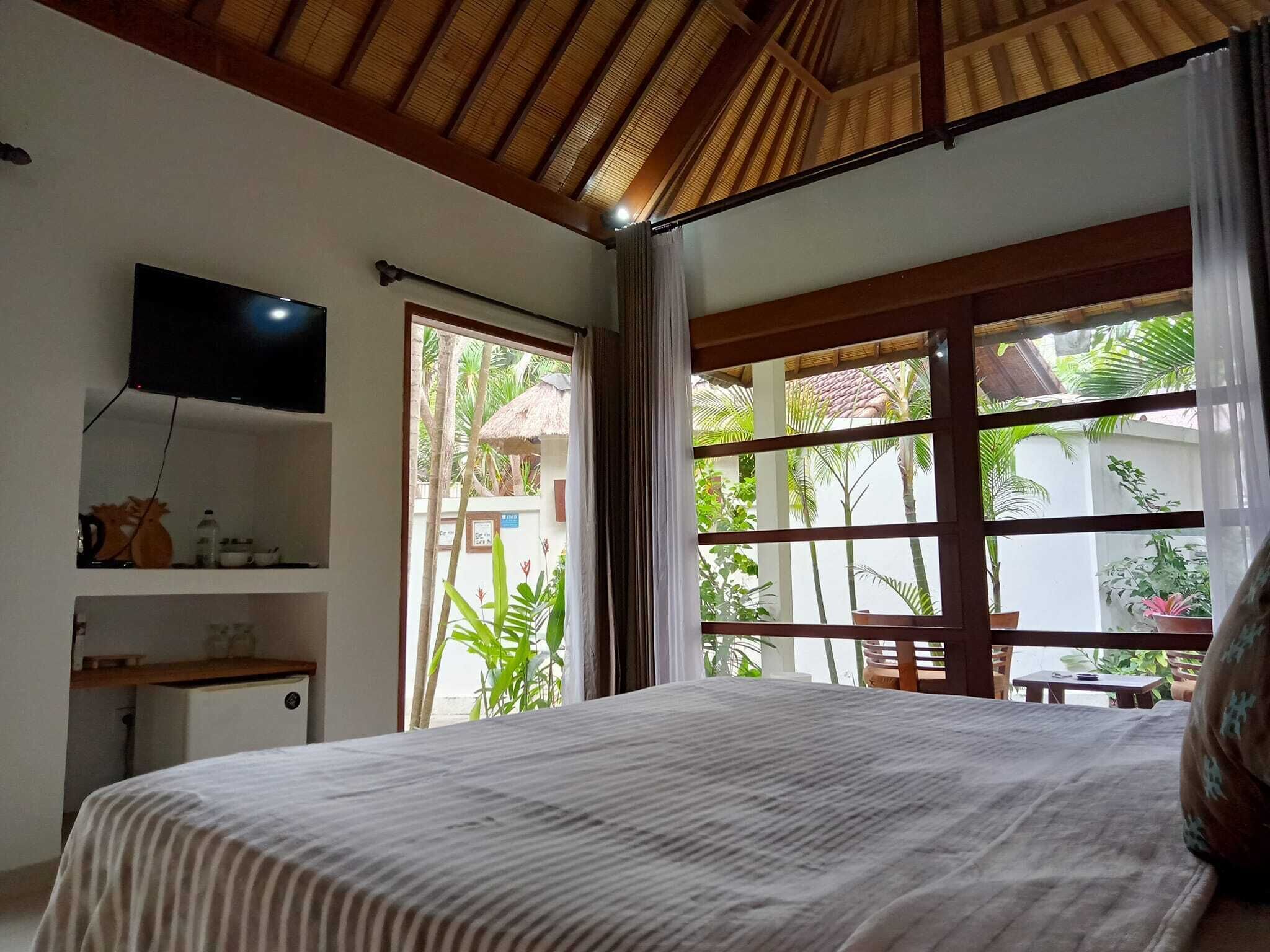 Un dormitorio con cama, televisor montado en la pared, repisa con objetos y ventana con cortinas.