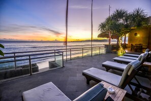 A terrace with lounge chairs, a table, and a view of the sea at sunset. - Balisanti Bungalows (Manggis)