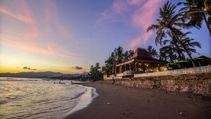 Une plage avec un bâtiment doté d’un mur en pierre et d’un toit, des palmiers et un coucher de soleil.