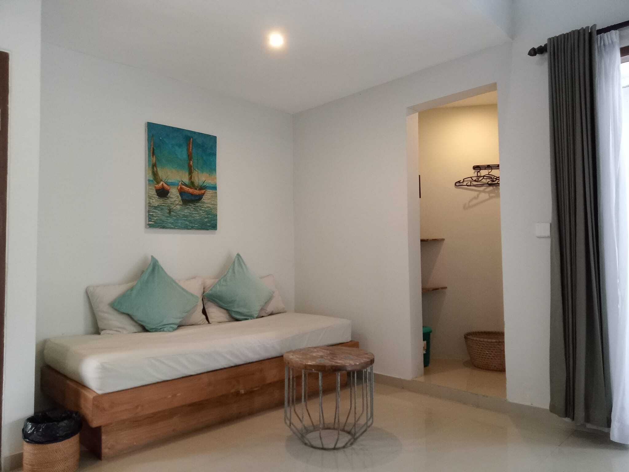 A room with a bed, a small stool, a painting of boats, and a doorway leading to another room.