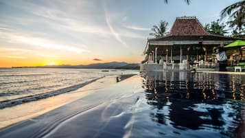 A beachfront resort with an infinity pool, a covered seating area, and a view of the sunset over the ocean.