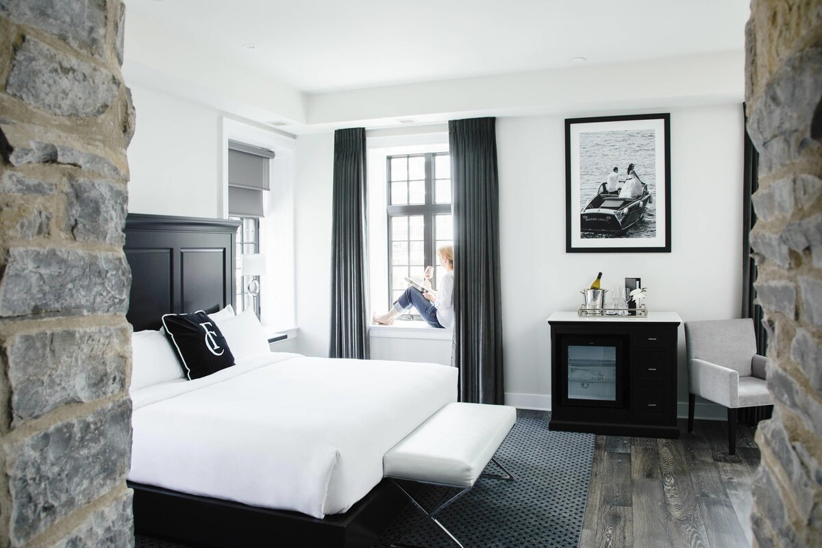 Una habitación de hotel con cama, escritorio, silla, ventana con cortinas e imagen enmarcada en la pared.