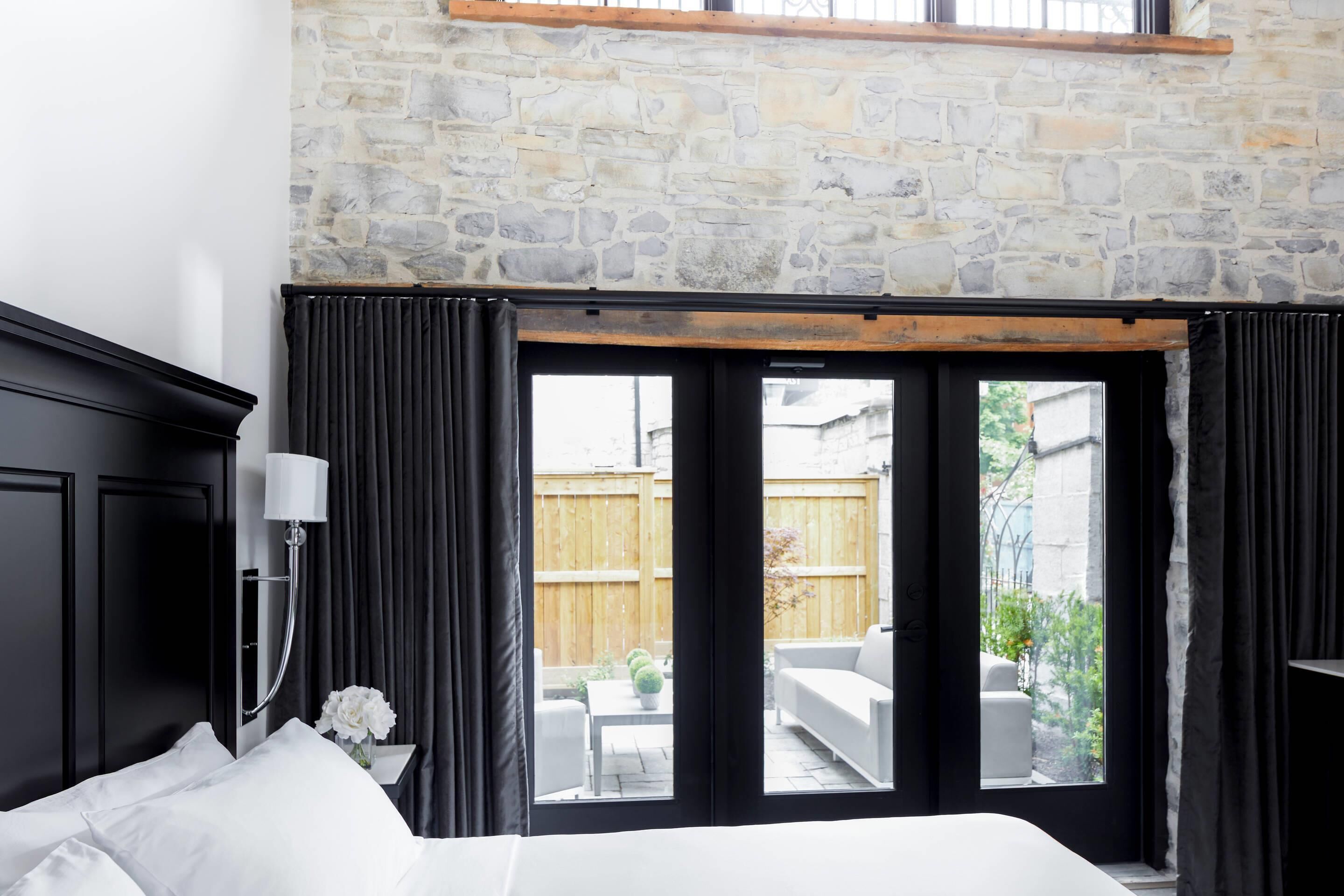 Quarto com parede de pedra, uma janela grande, cama, mesa de cabeceira, abajur e vista para o exterior.