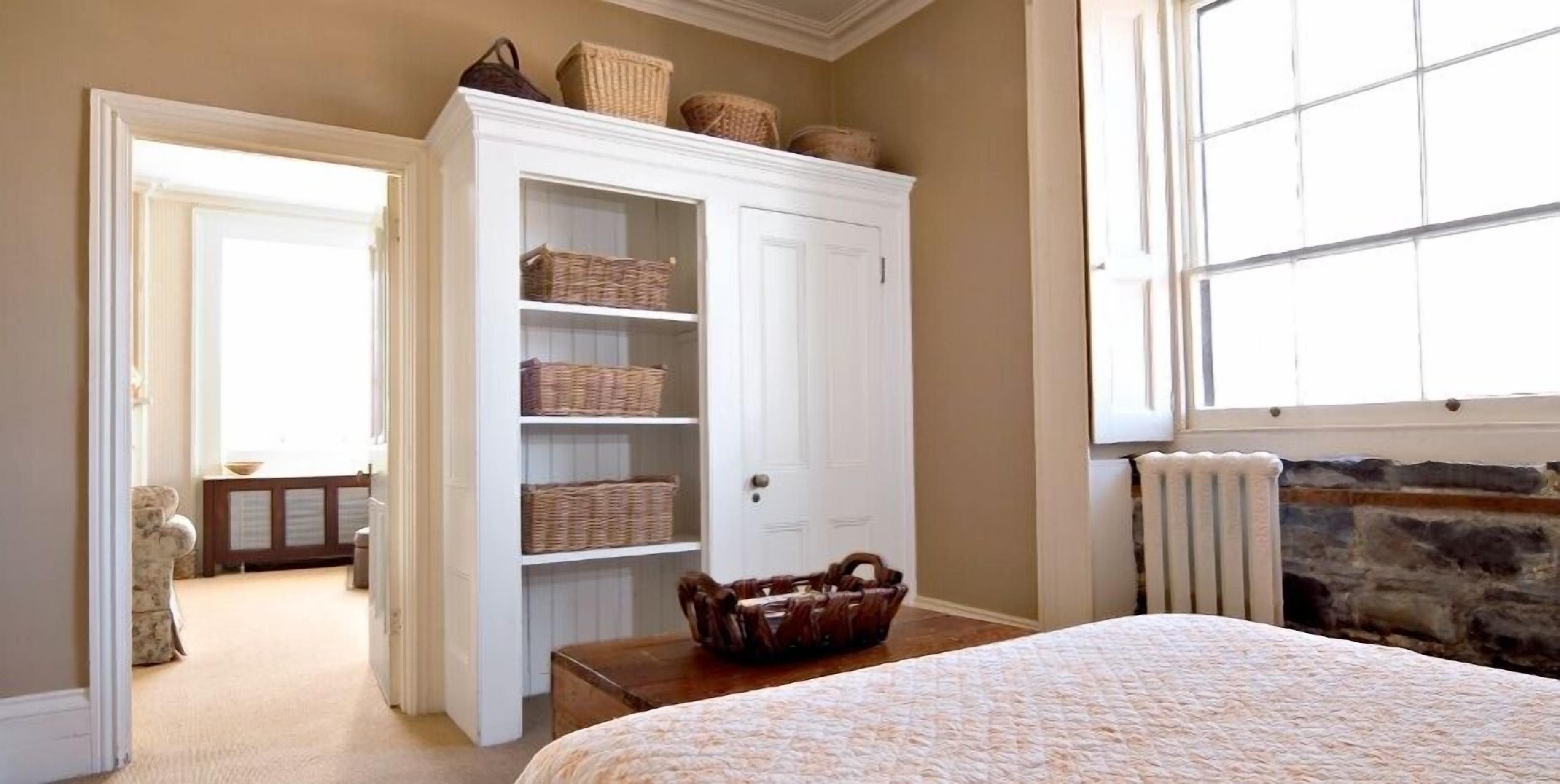 Une chambre à coucher avec un lit, une armoire blanche dotée de paniers en osier, une cheminée en pierre et une vue sur l’extérieur.