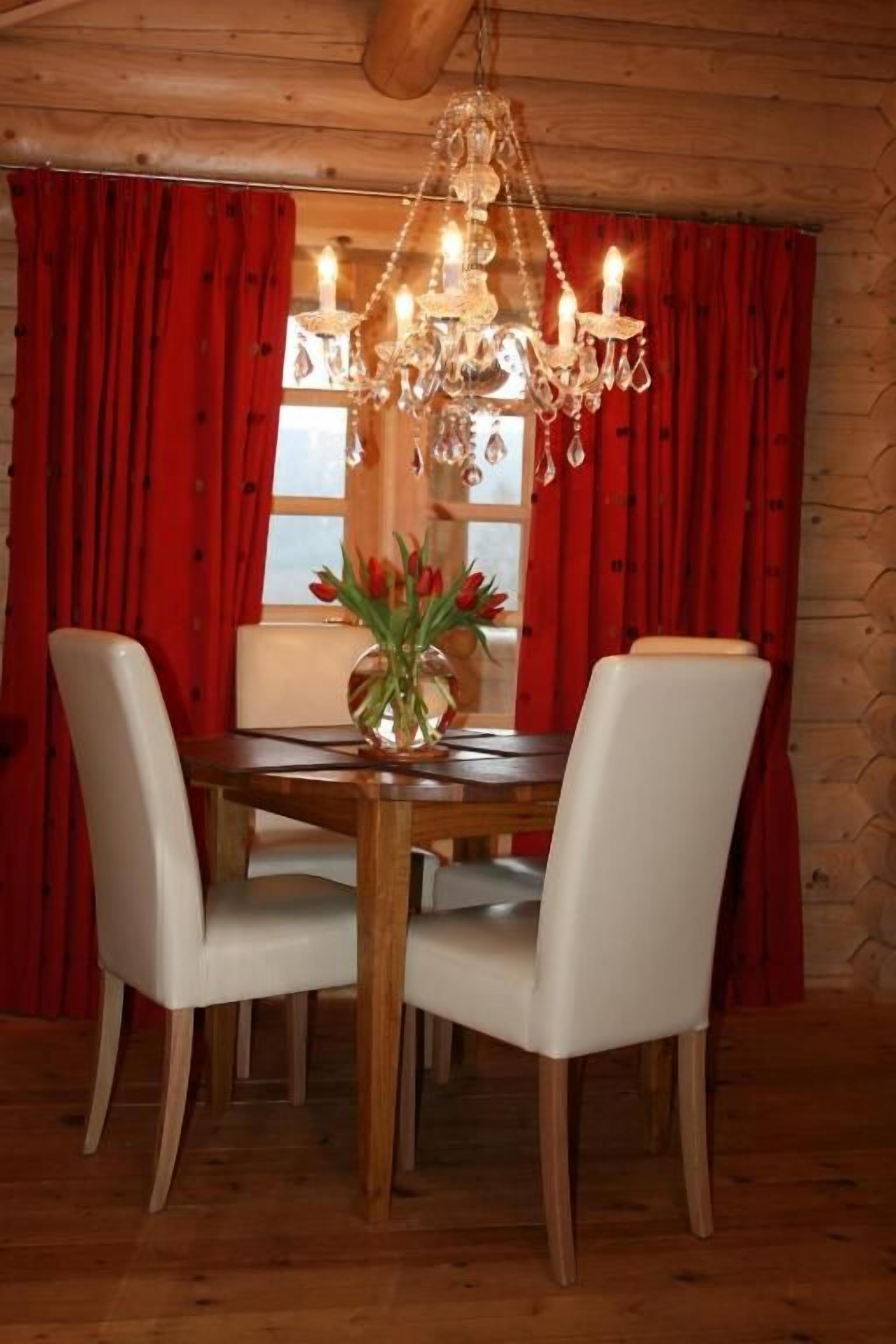 A dining area with a wooden table, white chairs, a chandelier, and red curtains.