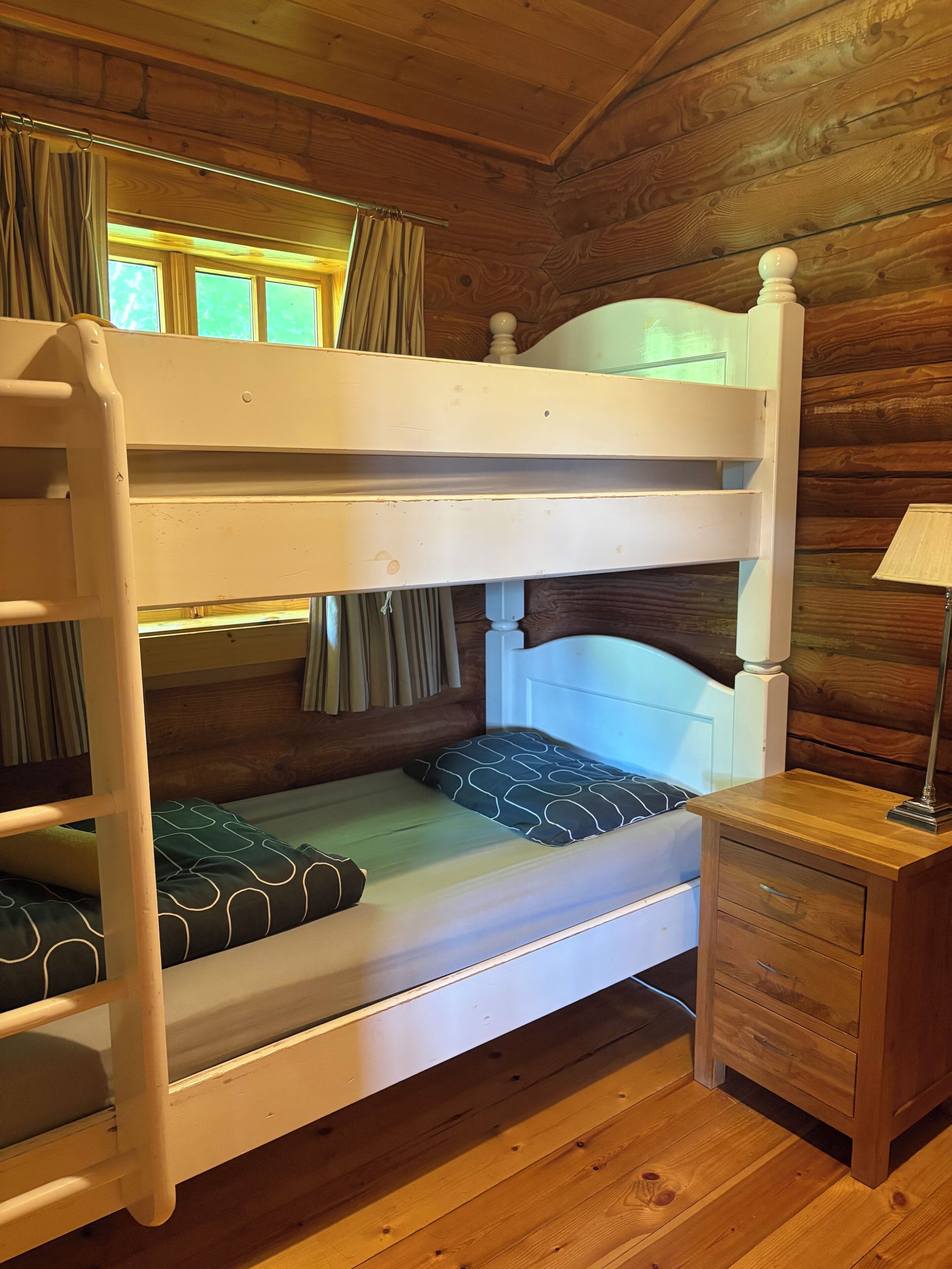 Een stapelbed met een houten hoofdbord, een nachtkastje met een lamp, en een raam met gordijnen.