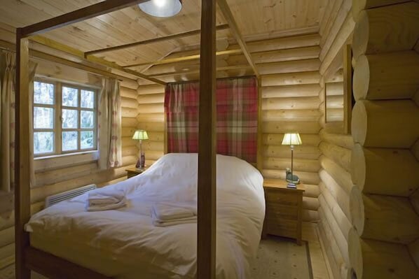 A log cabin bedroom with a four-poster bed, two bedside lamps, a window with curtains, and a plaid blanket on the bed.