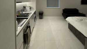 A modern, well-lit kitchen with a washing machine, a trash bin, and a dining area with a sofa and a TV. - Buckingham Apartments (High Wycombe)