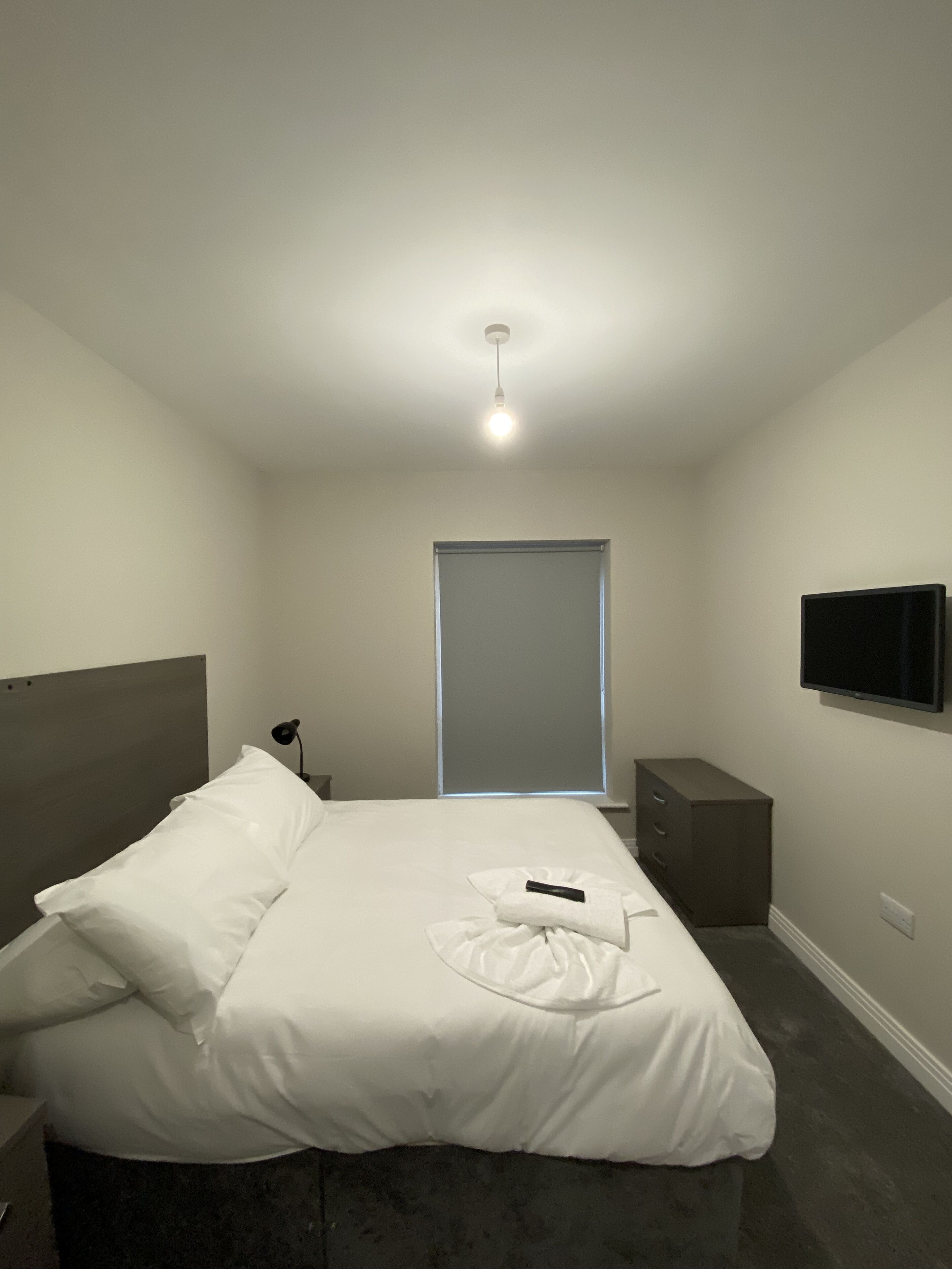 Une chambre d’hôtel minimaliste comprenant un lit, une table de chevet, un téléviseur fixé au mur et une fenêtre avec des stores.