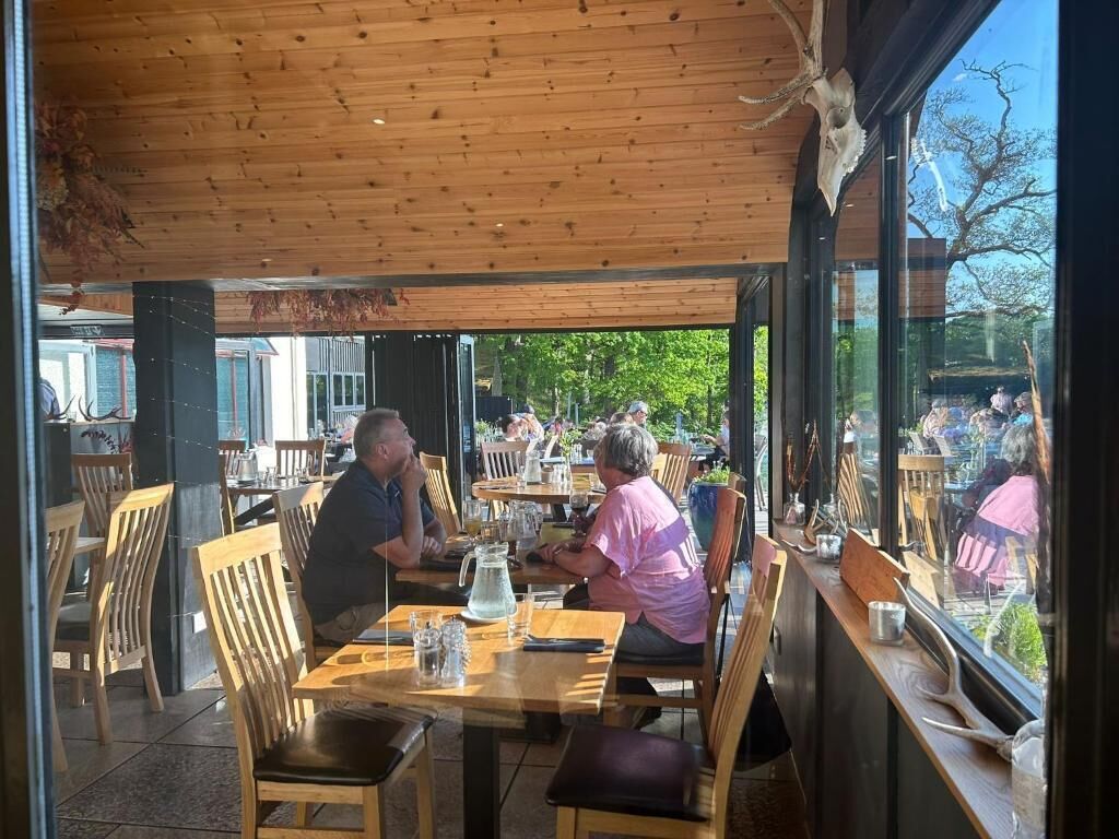 An outdoor dining area with wooden tables and chairs, large windows, and a view of the outdoors.
