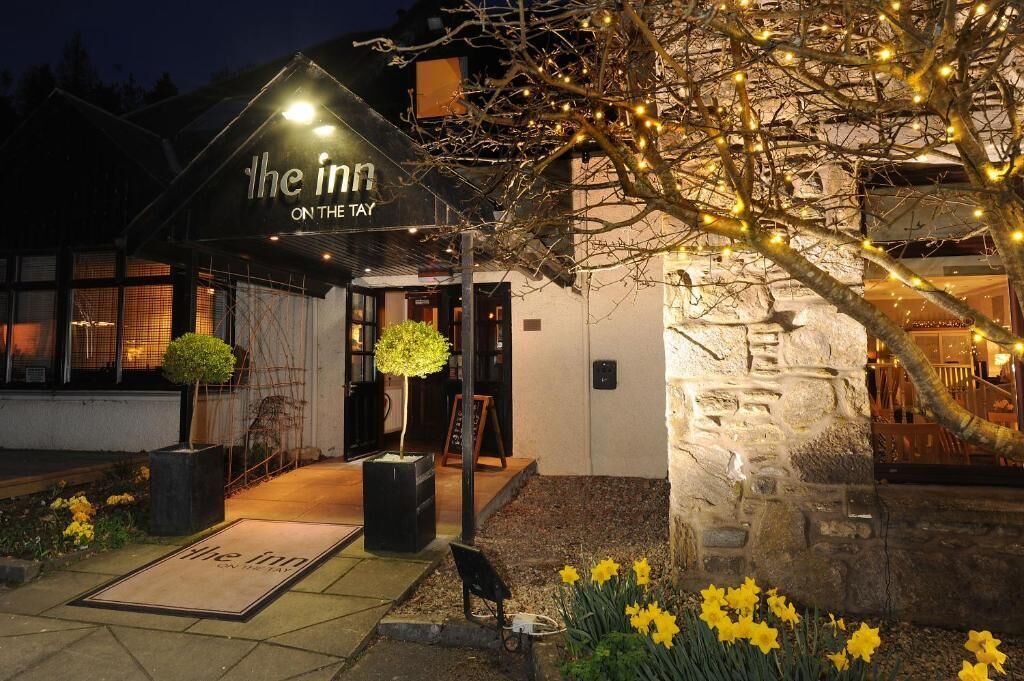 L’entrée d’un hôtel, avec une enseigne indiquant « The Inn on the Tay », et un paillasson portant le même texte. On y voit des plantes en pot et un arbre décoré de guirlandes lumineuses.