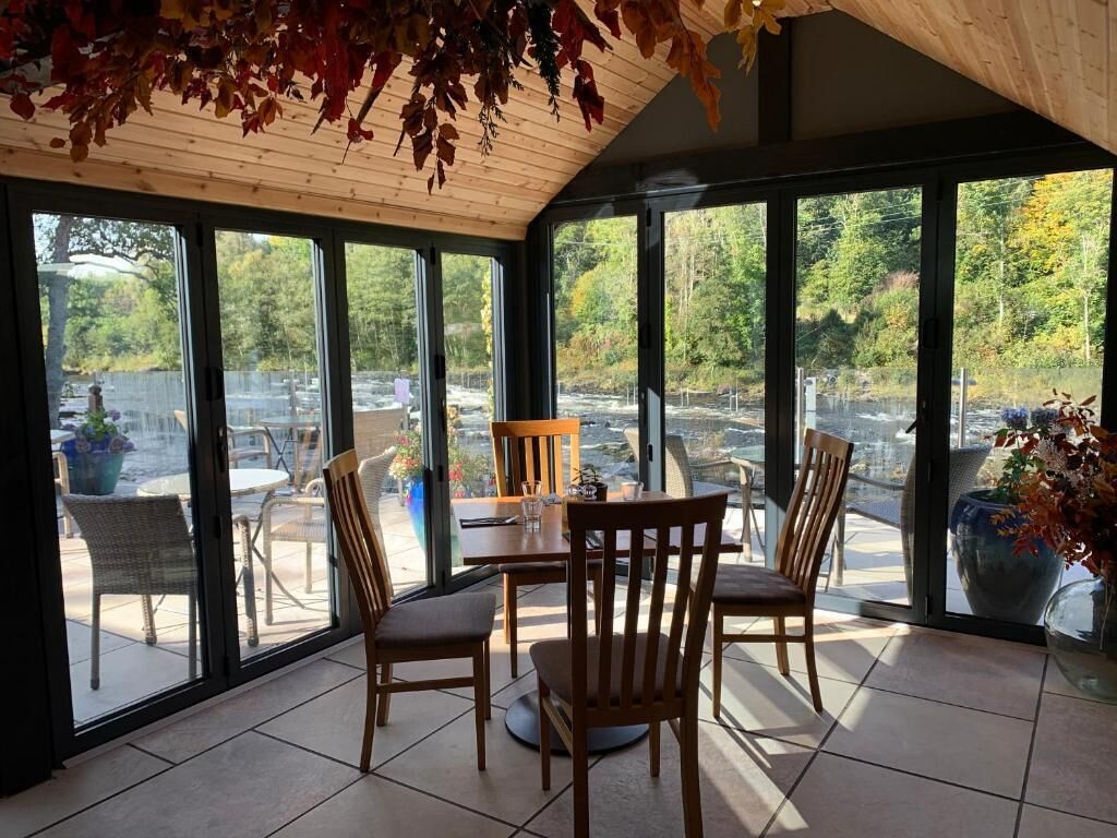 Une véranda avec une table à manger et des chaises, de grandes fenêtres et une vue sur un parking et des arbres.