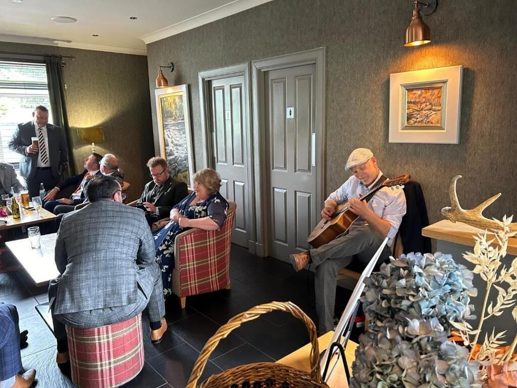 Un groupe de personnes est assis autour d’une table dans une pièce, et un musicien joue de la guitare.