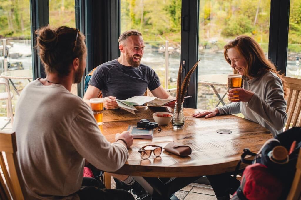 Three people seated around a round wooden table with a view of a river outside.