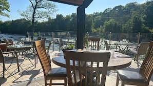 An outdoor patio with round tables and chairs, overlooking a body of water and surrounded by trees.