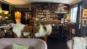 A bar area with a wooden counter, shelves of bottles, and a variety of glasses. There are several wooden chairs with fluffy white cushions.