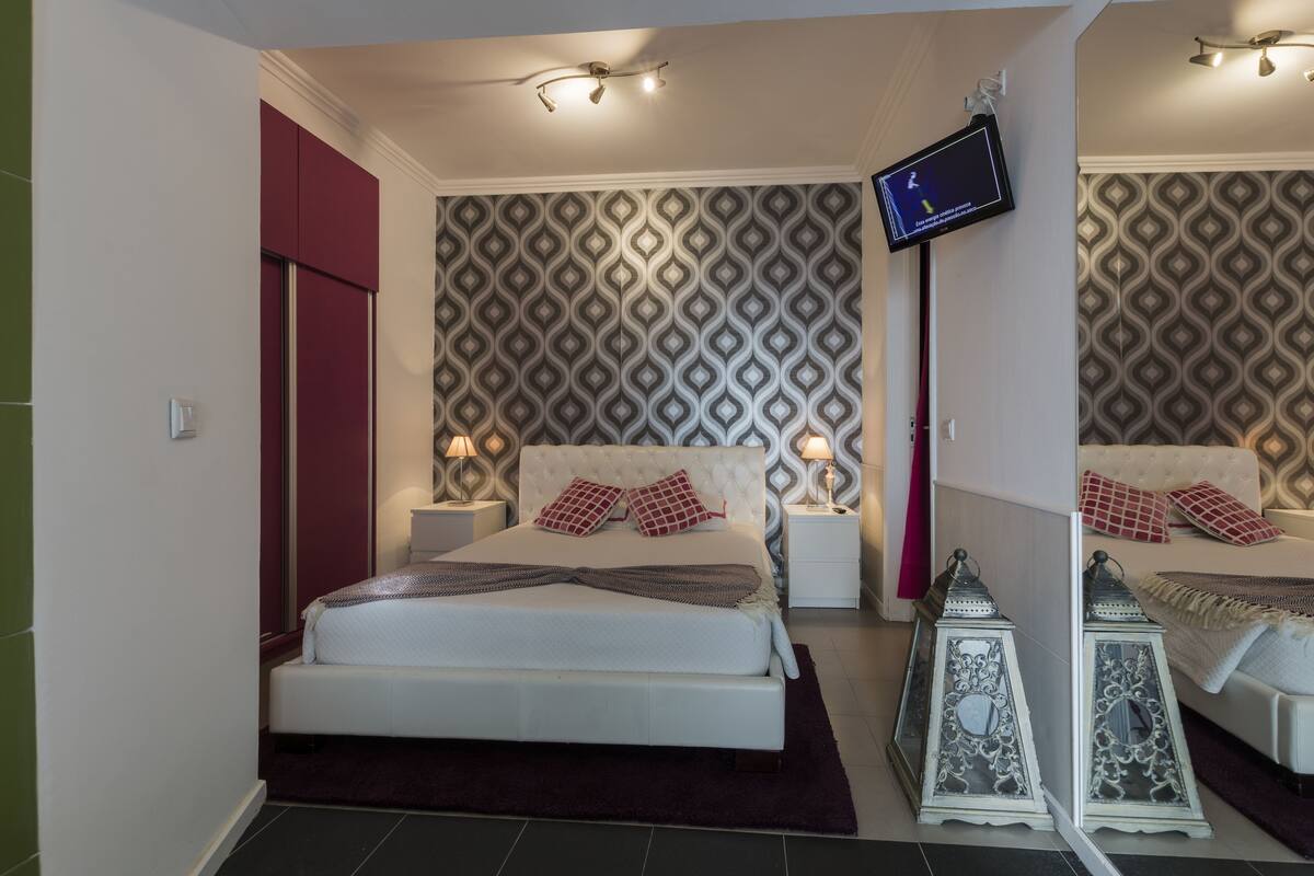 A modern bedroom with a large bed, a television mounted on the wall, patterned wallpaper, and decorative lanterns.