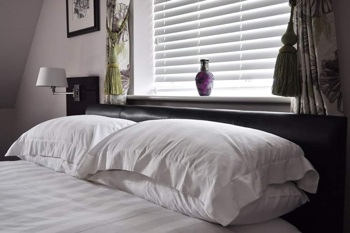 A neatly made bed with white linens, a black headboard, and a window with blinds and a vase.