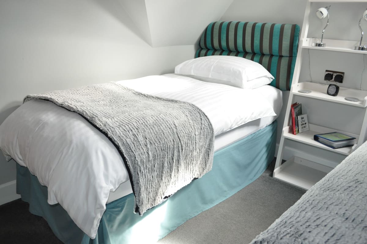 A single bed with a teal skirted base, white bedding, and a grey knitted throw. A white nightstand with a lamp and a small shelf holding books and a clock is beside the bed.